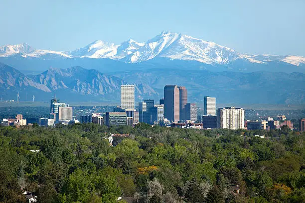 Snow covered Longs Peak, part of the Rocky Mountains stands tall in the background with green trees and the Downtown Denver skyscrapers as well as hotels, office buildings and apartment buildings filling the skyline.