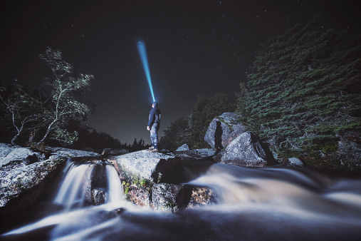 A man stands in the middle of a stream pointing his headlamp to Earth's North Celestial Pole, lying very near the bright star Polaris.  Long exposure.