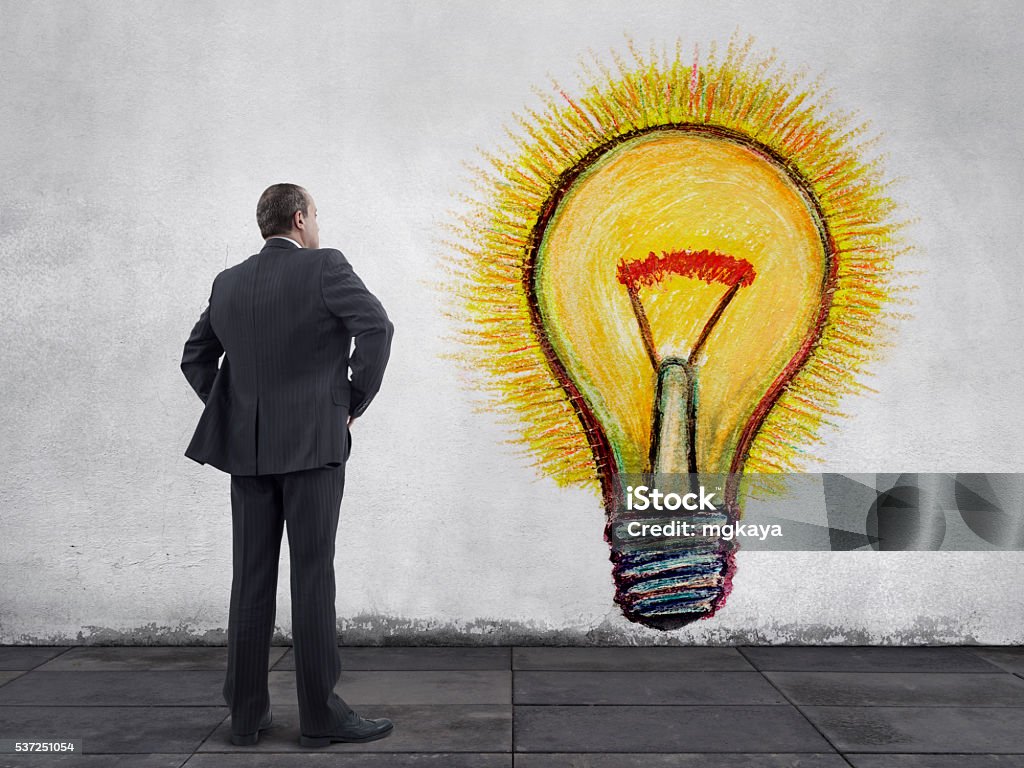 Businessman with Glowing Light Bulb Painting on the Wall Rear view of businessman standing on sidewalk and looking at the hand drawn glowing light bulb painting on the wall. Graffiti Stock Photo