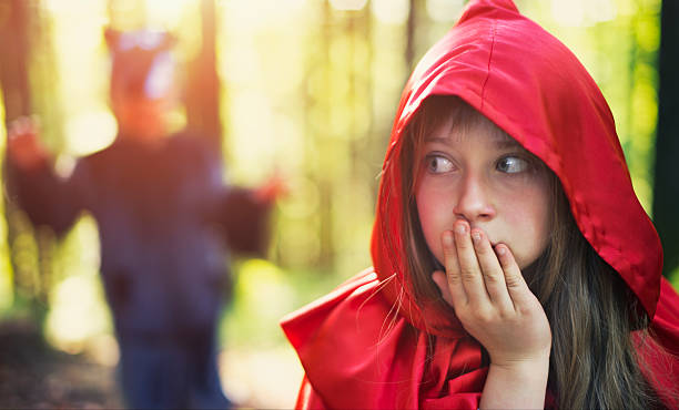 A little girl dressed as Little Red Riding Hood is wearing a red hood while walking through a burned out forest during Halloween. She has a worried expression on her face and is scared of the dark woods and shadows. Image taken in Utah, USA.