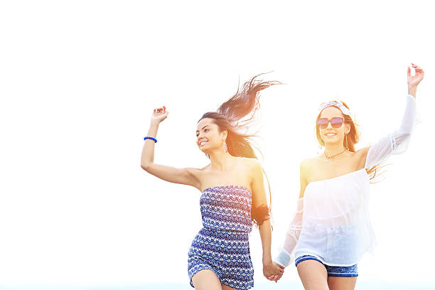 Two beautiful young women walking and having fun, jumping and dancing. Wears casual clothes, sunglasses and eyeglasses. Hands up.