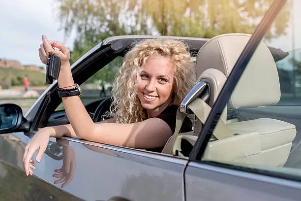 Young happy woman sitting in her new convertible car while holding car keys and looking at the camera.