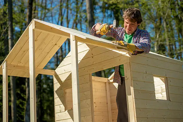 Young man using a mallet to fix a nail into a roof of a wooden playhouse for children in a DIY concept.