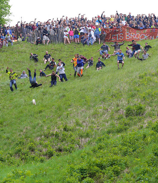 Brockworth, England - May 31, 2016: Entrants chasing the cheese at the 2016 ‘Cheese Rolling’ held at Cooper’s Hill, in the Cotswolds. Every spring bank holiday Monday on the 1 in 2 gradient (Yes 1 in 2!) ‘Coopers Hill’ in the depths of the beautiful Gloucestershire countryside, the slightly ‘More Crazy’ people of not only Gloucestershire but the world gather to pit themselves against ‘The Cheese’. The atmosphere at the event is a true joy with everyone helping each other scale the steep hill. In the true tradition of British stiff upper lip and community spirit a small band of enthusiasts from Brockworth and surrounding area take it upon themselves to organise, control and marshall the event