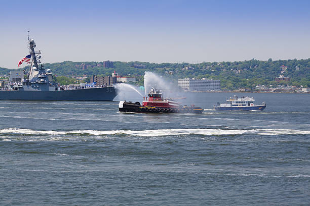 New York, NY, USA - May 25, 2016: NYC Fleet Week 2016, USS Bainbridge (DDG 96) guided missile destroyer entering New York Harbor. FDNY boat is shooting water from water cannons. Green trees of Staten Island are in background. US Navy sailors are lined up on the ship's deck under US Flag.