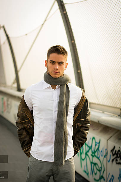 A pretty young man in a vintage stylish black leather jacket in a burgundy trendy golf dress and trendy black jeans stands in the studio against the background of the wall. Modern handsome guy.Fashion