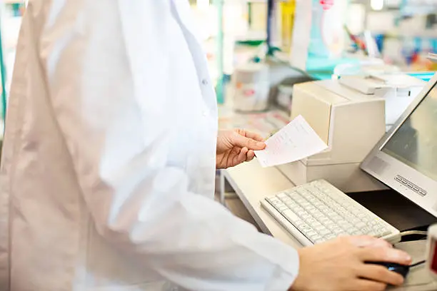 Closeup of pharmacist holding medication prescription and using computer at checkout counter to search medicine in the database