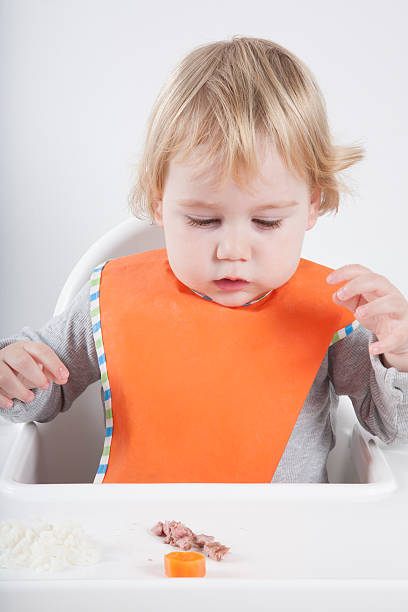 surprised baby eating blonde caucasian baby seventeen month age orange bib grey sweater eating meal in white high-chair surprised looking at carrot and tuna kids eating tuna stock pictures, royalty-free photos & images