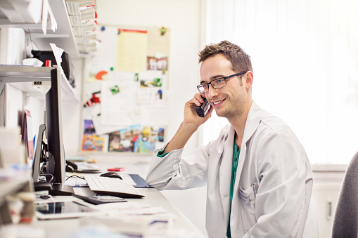 Happy male pharmacist sitting at his desk and talking on phone while working on computer at pharmacy