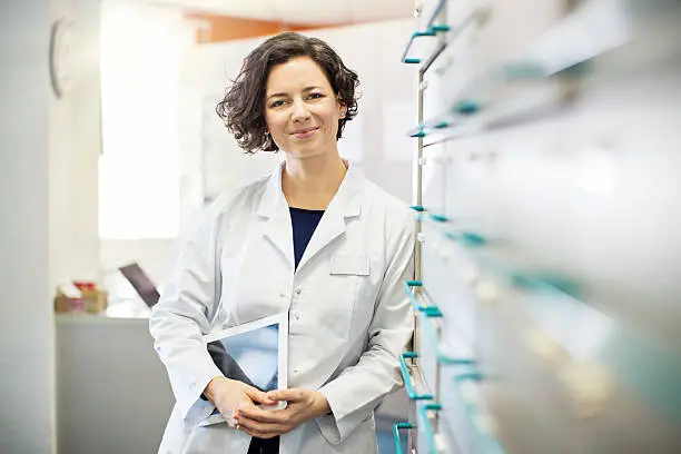 Portrait of confident young female pharmacist leaning to a medicine shelf with a digital tablet