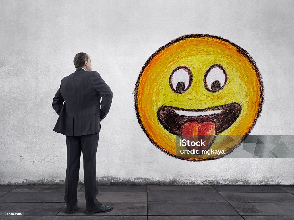 Businessman and Face with Stuck-Out Tongue Emoji Rear view of businessman standing on sidewalk and looking at the face with sticking out tongue emoticon hand drawn painting on the wall. Anthropomorphic Smiley Face Stock Photo