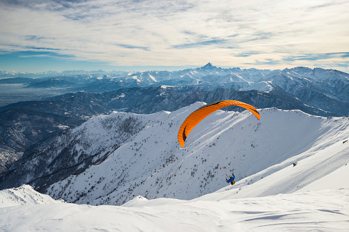 Paraglider flying on snowy slope with bright orange kite. Stunning background of the italian Alps in winter season. Shot taken in backlight, unrecognizable person.Paraglider flying over the mountains with bright orange kite. Stunning background of the italian Alps in winter season. Unrecognizable person.