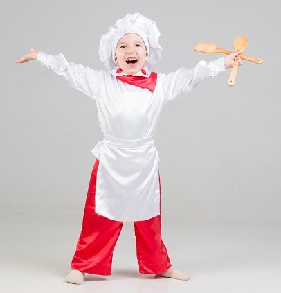 Laughing little boy dressed as a cook with chef cap on head. The little boy standing spread his arms wide open. The cute little boy is holding wooden spoon and spatula for cooking in hand. He is looking at the camera. Studio shooting on white background