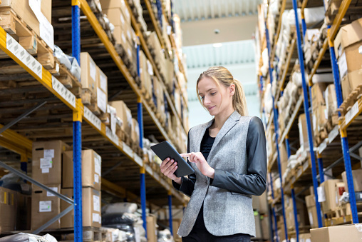 Portrait of a pensive young woman supervisor working on-line in warehouse. Young blond woman standing at distribution warehouse and wearing elegant suit. Industrial boss examining the stock. Large distribution storage in background with racks full of packages, boxes, pallets, crates ready to be delivered. Logistics, freight, shipping, receiving.