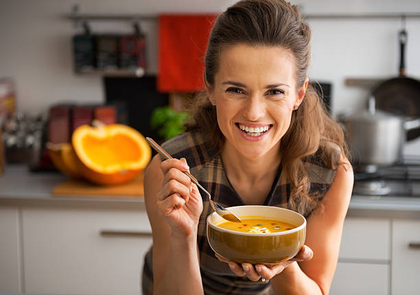 szczęśliwa młoda kobieta jedzenie zupa z dyni w kuchni - spoon heat pumpkin eating zdjęcia i obrazy z banku zdjęć