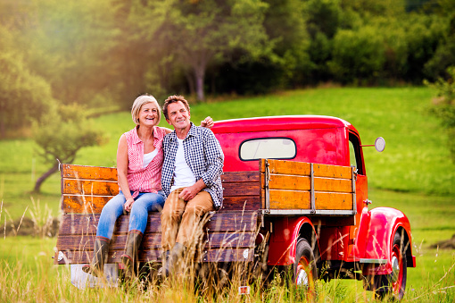 Senior couple sitting in back of vintage red pickup truck, green sunny nature