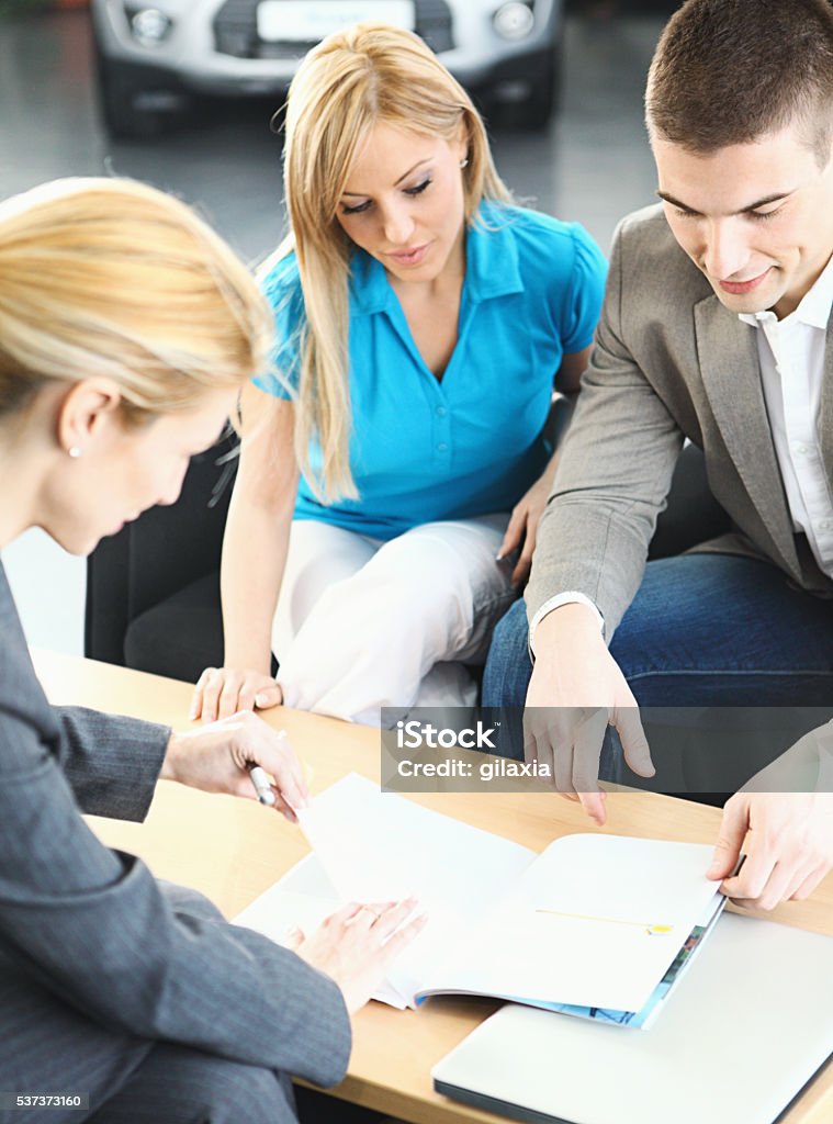 Couple buying new car. Car saleswoman showing vehicle brochure to a couple interested in buying a new car.They are in a meeting at a car dealership.Woman is wearing blue t-shirt and the man is in gray casual jacket. Blurry unrecognizable car in background. Car Stock Photo