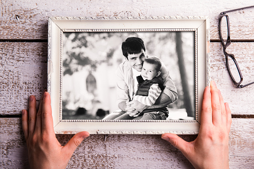 Fathers day composition. Hands of unrecognizable man holding black and white picture of father holding his little son. Studio shot on white wooden background.