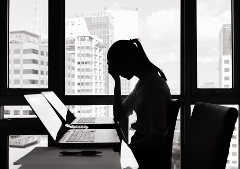 A photographic image of a stressed businesswoman sitting at a desk. The woman is seated in front of a window with a bright light source, rendering her image as a shadowed silhouette with indiscernible features. She is leaning forward with her elbows against the top of the desk and her hands clasped against her face, with her head slightly bent forward.
