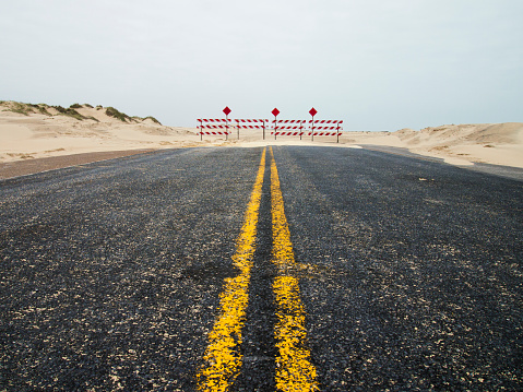 Last few feet of asphalt before red and white barriers indicate the end of the road.  Sand dunes line the road edges.