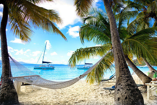 Serene hammock on a quiet beach on a tropical island with sailboat in the bay.