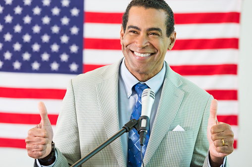 Happy African American politician gives to thumbs up at the podium. He is wearing a suit jacket and a blue tie.