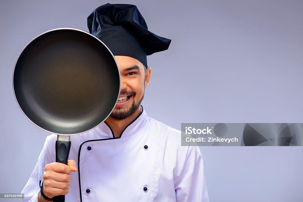 Portrait of young handsome cook He is a best chef ever. Closeup portrait of handsome cook covering a half of his face with a frying pan and smiling while standing against grey background with copy space 2015 Stock Photo