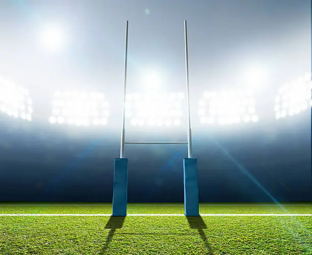 A rugby stadium with rugby posts on a marked green grass pitch at night under illuminated floodlights