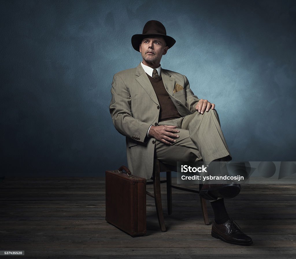 Fashionable retro 1940 business man with hat sitting on chair. Fashionable retro 1940 business man with hat sitting on chair in room. Men Stock Photo