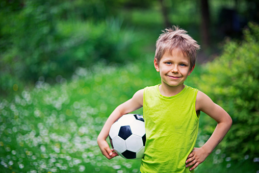 Little boy is playing football in the garden. The boy aged 6 is holding the football and smiling at the camera. Spring garden in the background.