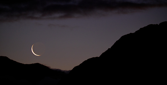 The sun has set on the Southern Alps in the Arthur's Pass National Park, on New Zealand's South Island. A new moon appears as the thinnest crescent, contrasting with the silhouetted mass of the mountains.