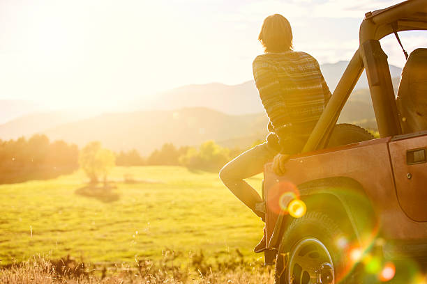 Man sitting on back of SUV parked at field Rear view of young man sitting on back of SUV parked at field sunny day stock pictures, royalty-free photos & images