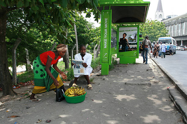 Dar es Salaam, Tanzania - February 21, 2008: Dar es Salaam, Tanzania - February 21, 2008: Black African woman sells bananas next to the green bus stop, close to the black African man reading a newspaper.
