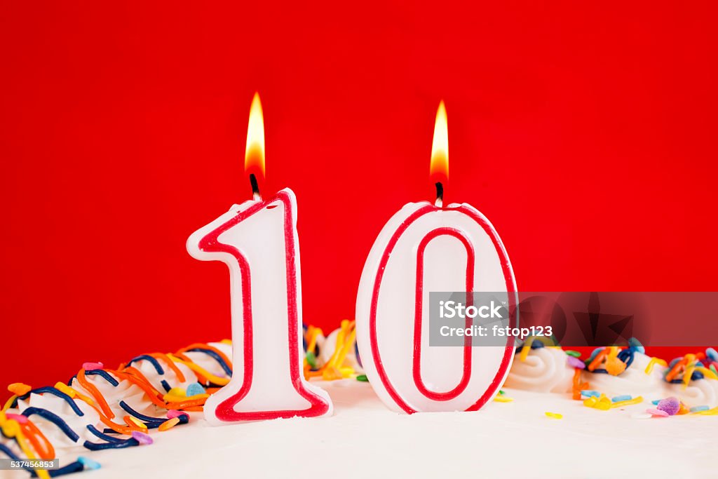 Decorated birthday cake with number 10 lit candles.  Red background. Decorated birthday cake with number 10 burning candles.  Red background.  White icing with multi-colored confetti. No people. 10th anniversary or 10th birthday celebration.  Close up. 2015 Stock Photo