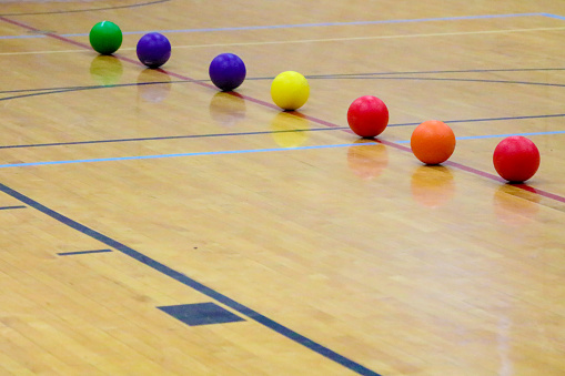 Dodgeballs lined up in the center of a gymnasium waiting for the start of the game.  The seven balls are red, orange, yellow, blue and green.  Dodgeball is an indoor team sport.