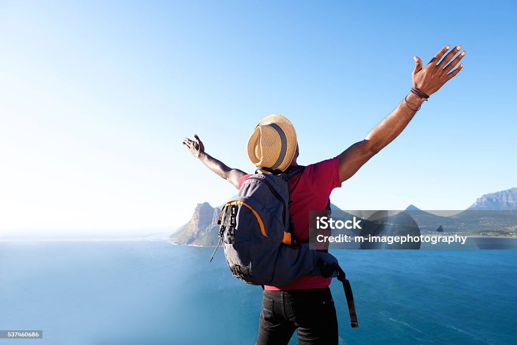 Young african guy on vacation Rear view portrait of young african guy with hat and backpack standing outdoors with arms spread open Travel Stock Photo