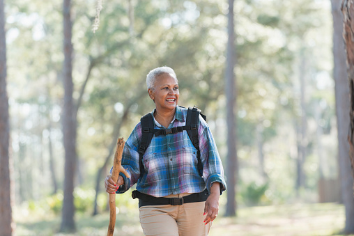 A senior African American woman carrying a backpack and walking stick, hiking through the woods.