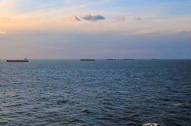 Five cargo ships ride at anchor in the North Sea off the coast of Belgium as the sun sets. Many ships can be seen waiting for a call to pick up cargo or being used as floating oil tanks