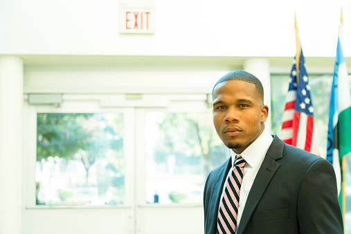 African descent, male attorney or politician in American courthouse building or congressional offices.  Or principal, professor in school building.  Red, white, and blue USA flag in background. He wears a full suit and tie and looks seriously at camera. Handsome, confident, successful.