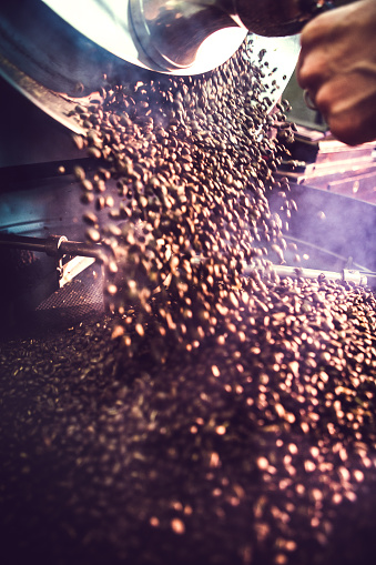 The process of roasting a batch of high quality single origin coffee beans in a large industrial roaster.  A mans hand is visible releasing the roasted beans into the cooling cycle.  Smoke rises from the hot beans.  Vertical with slight motion blur and copy space.  