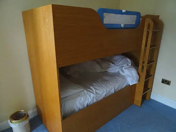 Photo showing a wooden bunk-bed in a small bedroom with a bed guard-rail / bedrail on the top bunk, to stop children falling out of the bed and ultimately make them feel more secure whilst sleeping.  A wooden ladder connects the top bunk.