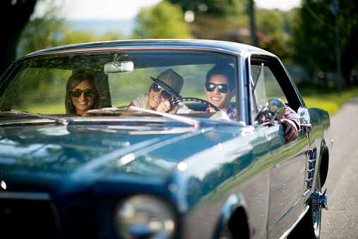 A multi-ethnic group of college age students are taking a road trip together on their spring break. They are driving a classic car on a sunny day.
