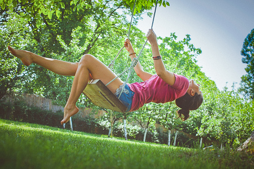 A happy young woman laughing on a bright sunny day while having fun on a swing in the garden. She grins widely and kicks her legs in the air as she reaches the apex of her swing. The image is taken from below as she swings, and there are leafy trees and a clear blue sky in the background.