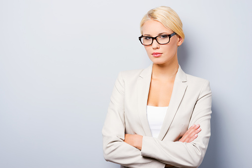 Confident young businesswoman keeping arms crossed and looking at camera while standing against grey background