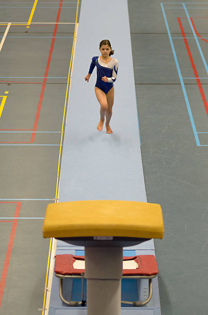 A small gymnast girl makes performance with gymnastic ball on a large professional stage. She is wearing a professional swimsuit for gymnastics. Behind her is a stadium with spectators.