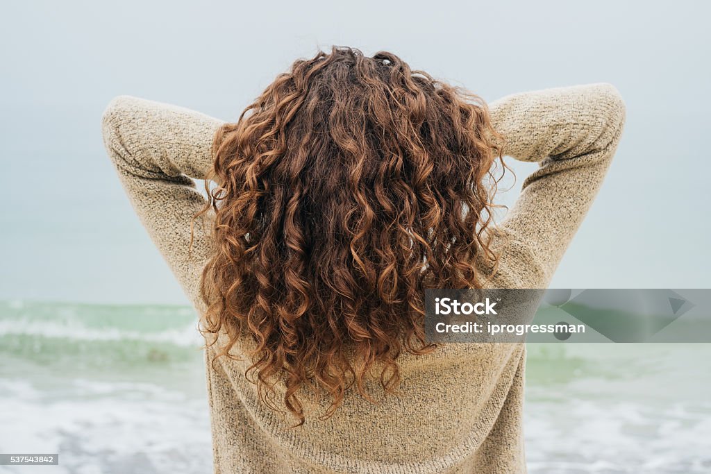 Curly brunette in a sweater on the coast Curly brunette in a sweater on the coast faces the sea and straightens hair, view from the back Curly Hair Stock Photo