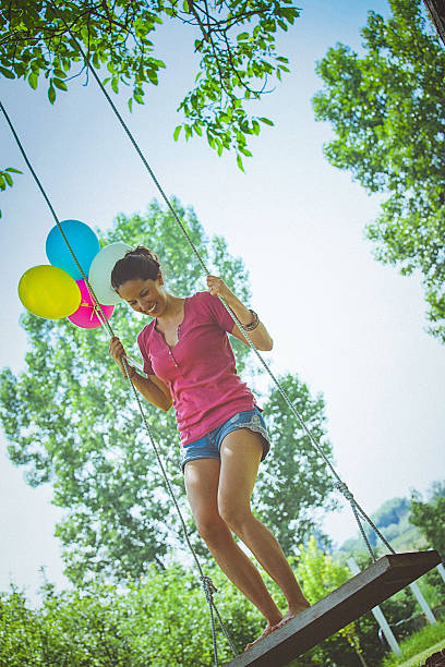 Happy young woman smiles while having fun on a swing in the garden, on a bright sunny day. She holds a bunch of colourful balloons, while standing on a swing. The image is taken from the low angle, and there are green leafy trees and a clear blue sky in the background.