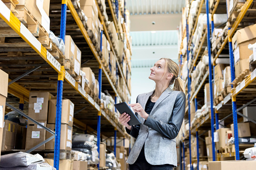 Horizontal color image of a pensive young woman supervisor with digital tablet in hand looking up in warehouse. Young blond woman standing at distribution warehouse and wearing elegant suit. Industrial boss examining the stock. Logistic worker working in a large distribution warehouse. Large distribution storage in background with racks full of packages, boxes, pallets, crates ready to be delivered. Logistics, freight, shipping, receiving.