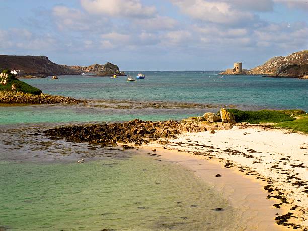 Cornwall England beach, Cromwell’s Castle, Tresco, Isles of Scilly Turquoise water laps a sandy beach on Tresco island in the Isles of Scilly, an archipelago off the coast of Cornwall, England. Cromwell's Castle towers over the coast in the distance. tresco stock pictures, royalty-free photos & images