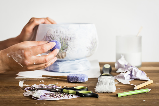 Decoupage artist workshop: scissors, sponge, paintbrush, pencils and paint. Hands of a hobbyist decorating a vase with lavender pattern.